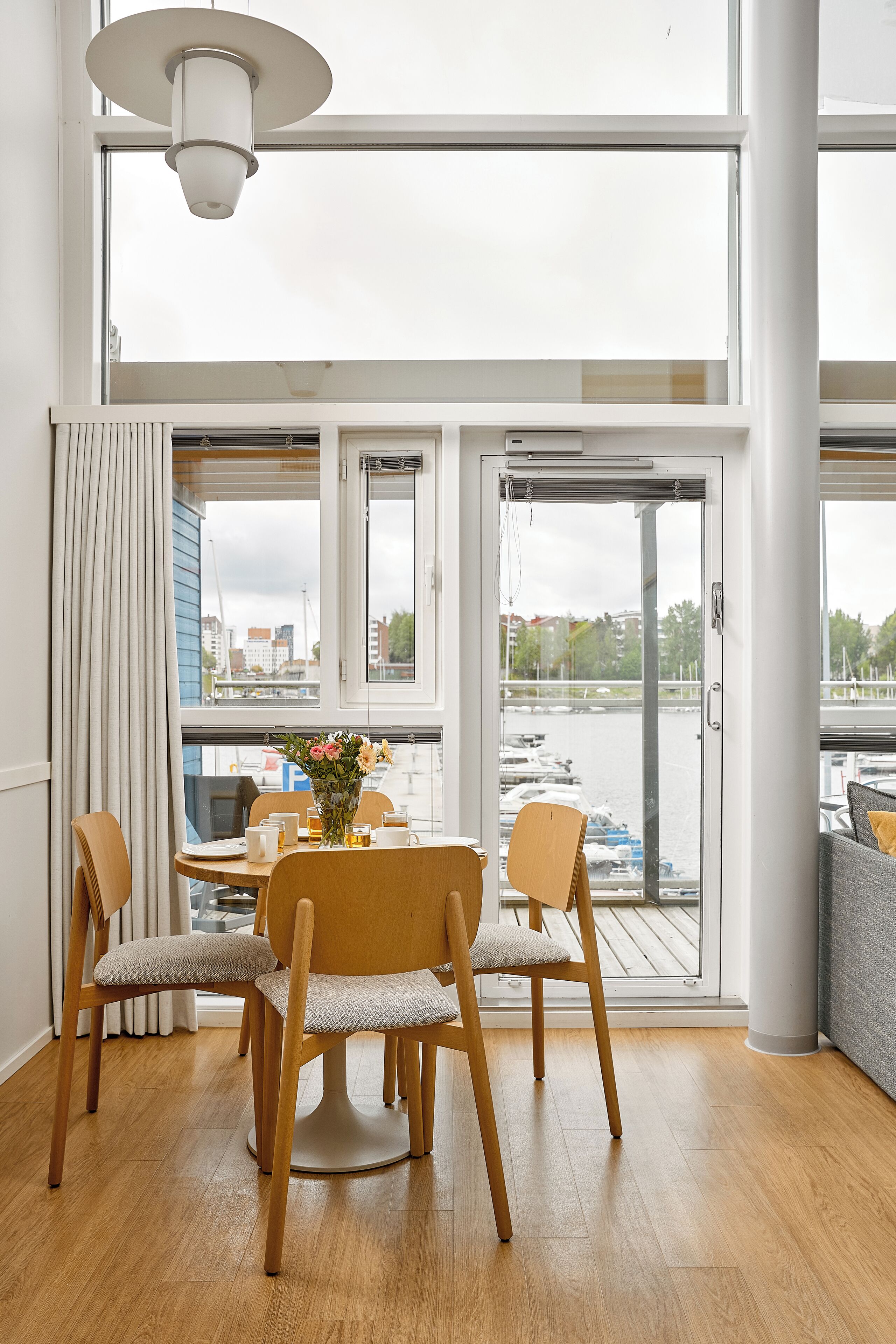 Un coin repas avec une table et des chaises, un vase rempli de fleurs et une vue sur une étendue d’eau, visible à travers de grandes fenêtres.