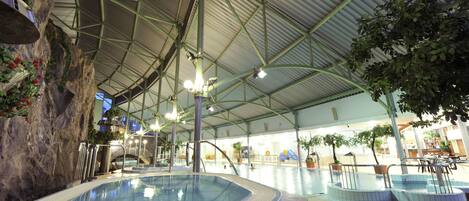 An indoor swimming pool with multiple hot tubs, surrounded by plants and a high ceiling structure.