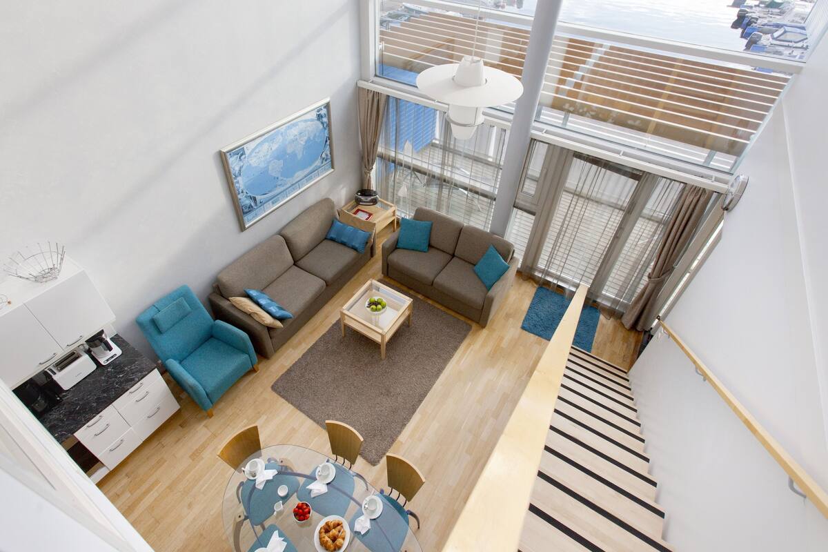 A modern living room with a sofa, chairs, a coffee table, and a dining table. There is a large window with a view of a marina.