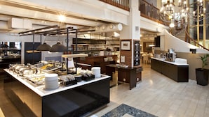 Um hall de hotel espaçoso, com teto alto, balcão de recepção central, espaço para buffet e escada que dá acesso a um andar superior.