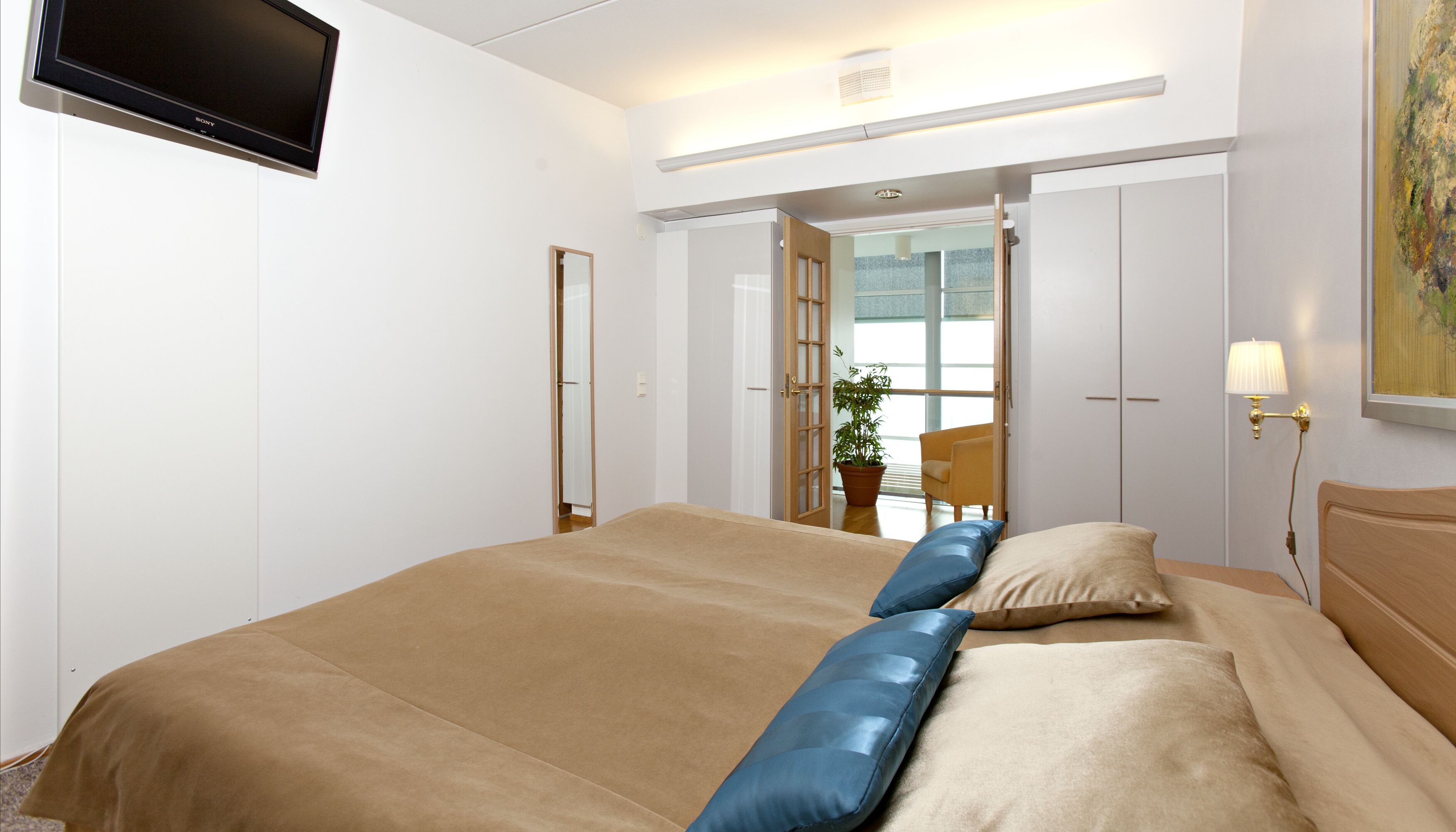 A hotel room with a bed, a TV mounted on the wall, a door leading to a balcony, and a lamp on the nightstand.