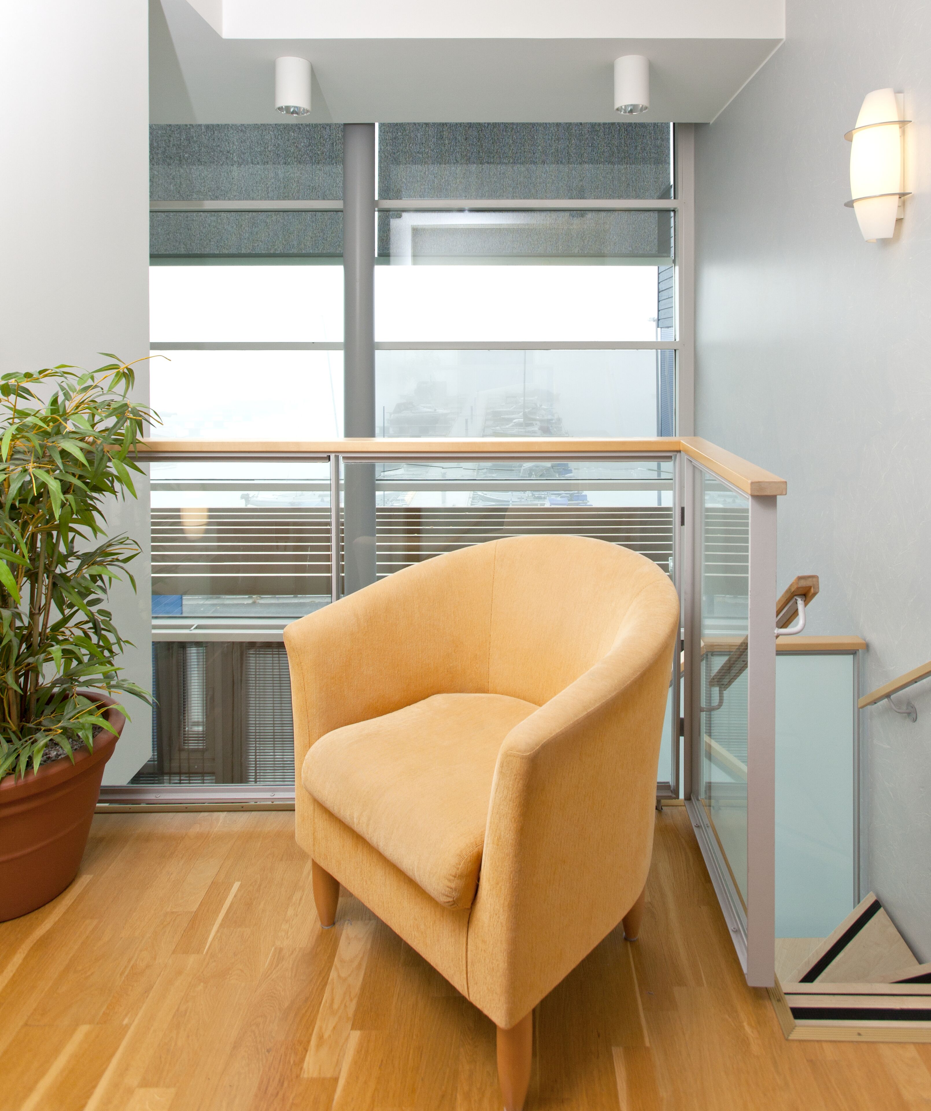 Una habitación moderna con piso de madera, un sillón color beige, una planta en maceta y barandilla de vidrio.