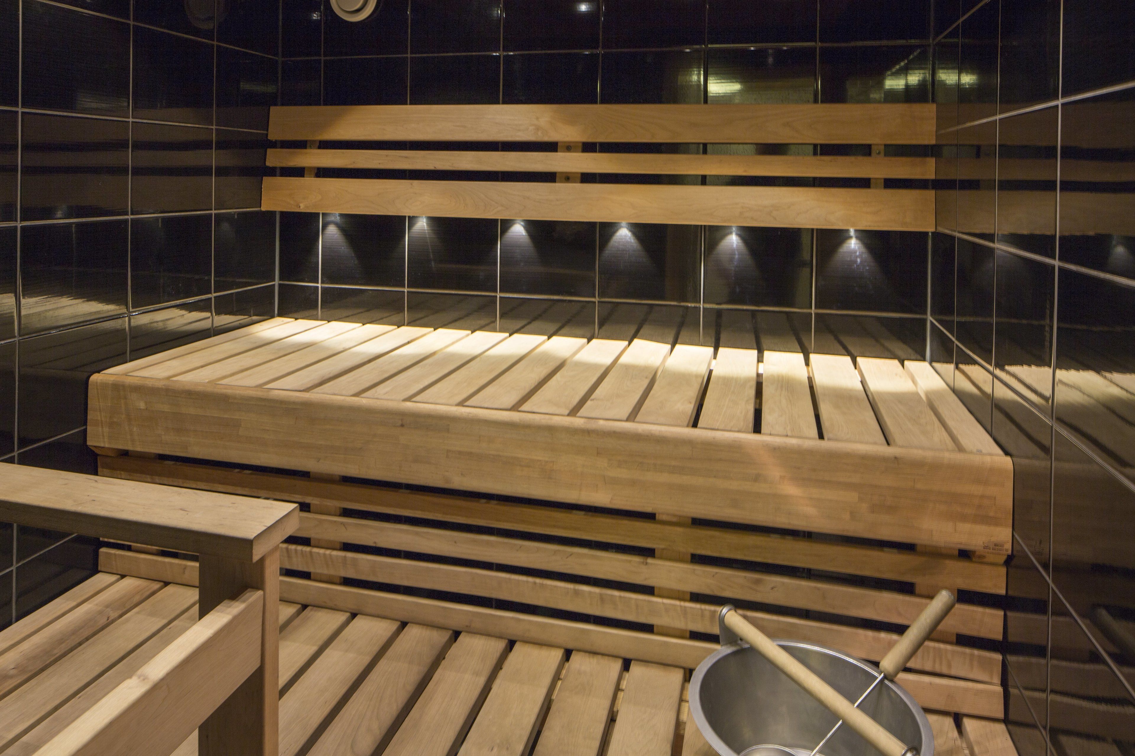 A wooden bench with a metal bucket and a wooden stool in a tiled room with a bench in the background.