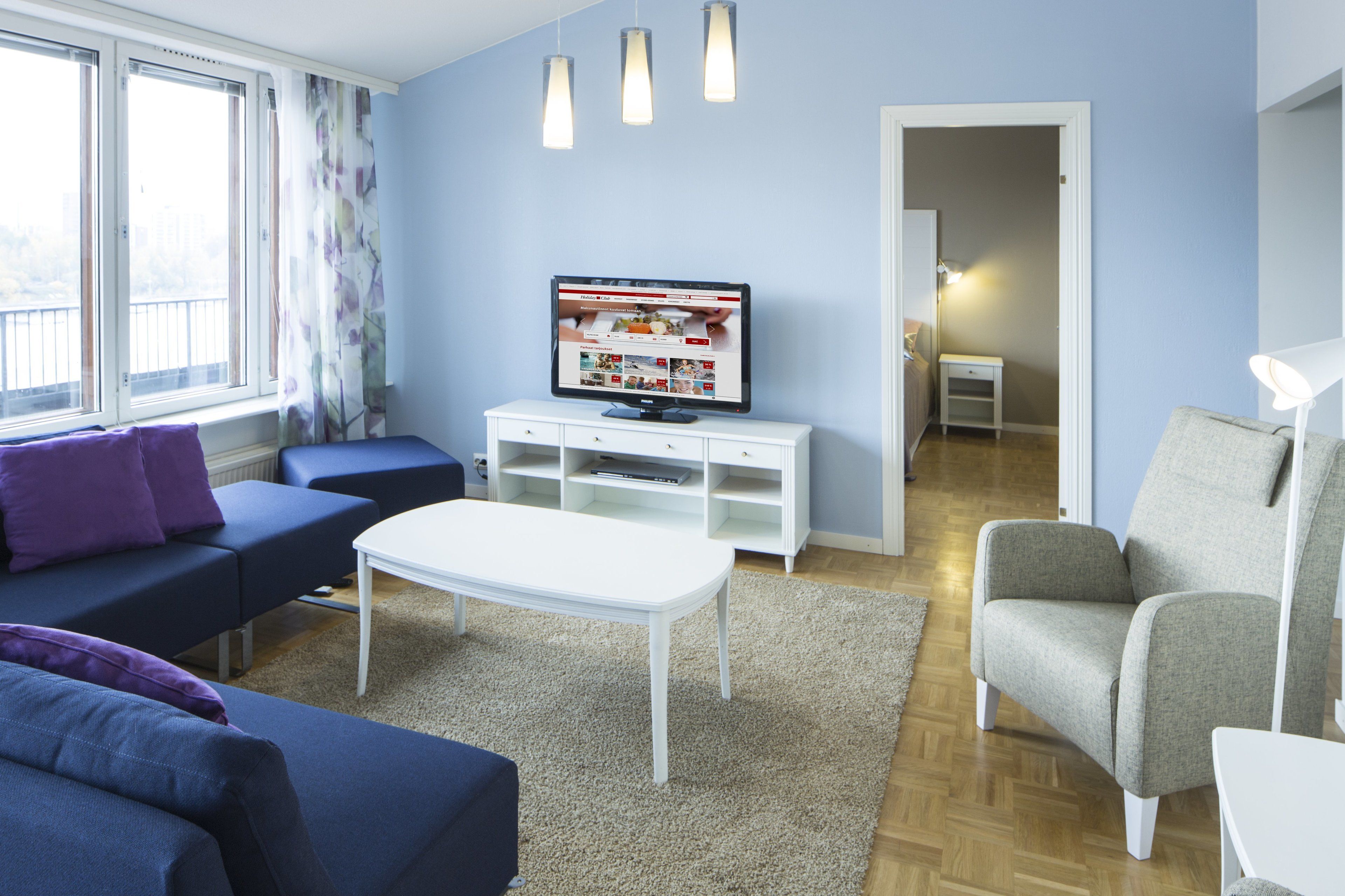 A living room with a sofa, armchair, and a coffee table. There is a TV on a stand and a window with curtains.