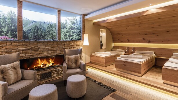 A modern living room with a stone fireplace, wooden ceiling, and a view of a forested mountain. - Hotel Chesa Monte (Fiss)