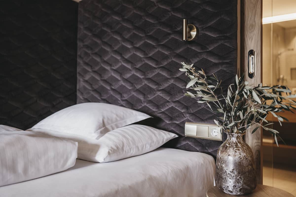Un lit bien fait, avec du linge de lit blanc, un mur sombre à la texture intéressante derrière, et un vase avec des plantes vertes sur une table en bois placée à côté du lit.