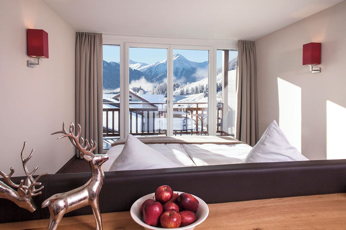 Une chambre d’hôtel avec un lit, une vue sur les montagnes, un balcon et une table sur laquelle se trouvent des pommes et une sculpture de cerf.