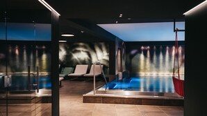 A modern indoor pool area with clear glass walls, a hot tub, and a seating area with chairs. - Hotel Chesa Monte (Fiss)
