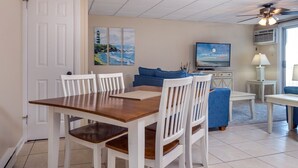 A dining area with a wooden table and white chairs, a blue sofa, a television on a stand, and a ceiling fan. - Oceanfront condos with 3 night minimum rentals and linens included! (Ocean City)