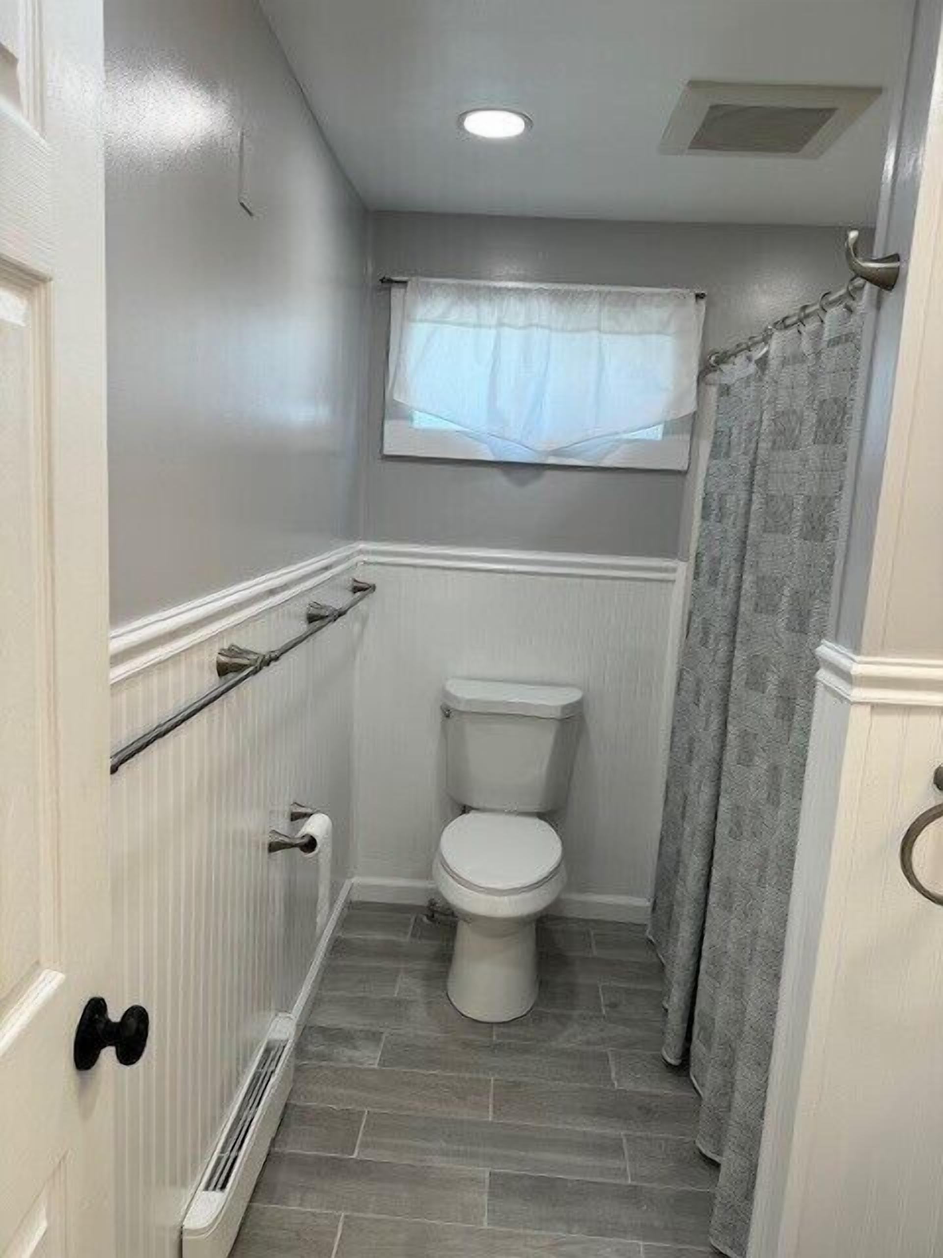 A small bathroom with a toilet, a window with a sheer curtain, and a shower curtain.