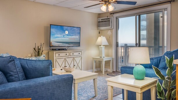 A living room with a television, blue sofas, a ceiling fan, and a view through a sliding glass door. - Oceanfront condos with 3 night minimum rentals and linens included! (Ocean City)