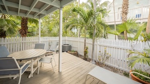 Une terrasse couverte avec une table et des chaises en bois, entourée d’une clôture blanche en piquets et de diverses plantes.