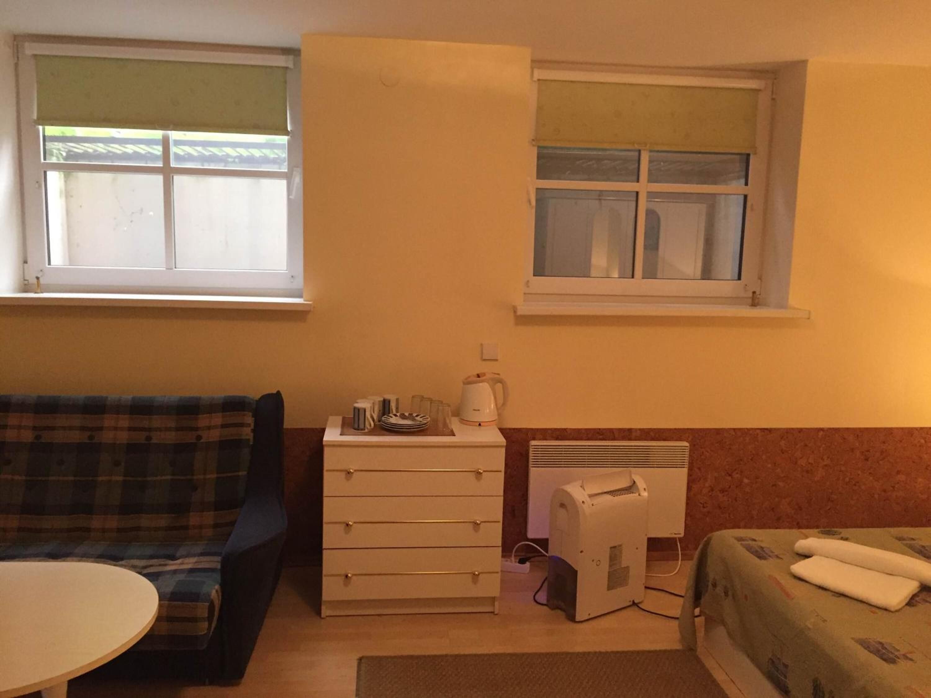 A small, cozy room with a sofa, a table, a dresser, and a bed. There are two windows providing natural light.