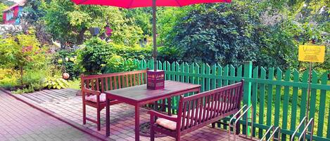 A patio with a table and two chairs under a red umbrella, surrounded by a green fence and lush greenery.
