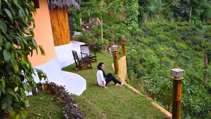 Una persona sentada en una ladera cubierta de hierba, con una construcción de techo de paja y sillas cerca.