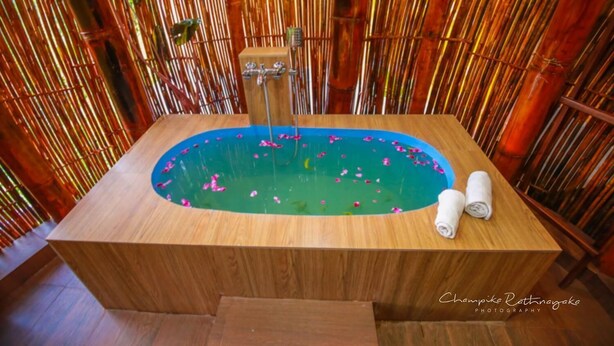 Una bañera de madera con interior azul, rodeada de paredes de bambú y dos toallas enrolladas a un lado.