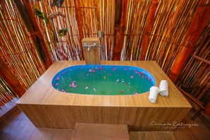 A wooden bathtub with a blue interior, surrounded by bamboo walls, and two rolled towels on the side. - Tea Cabins (Ella)