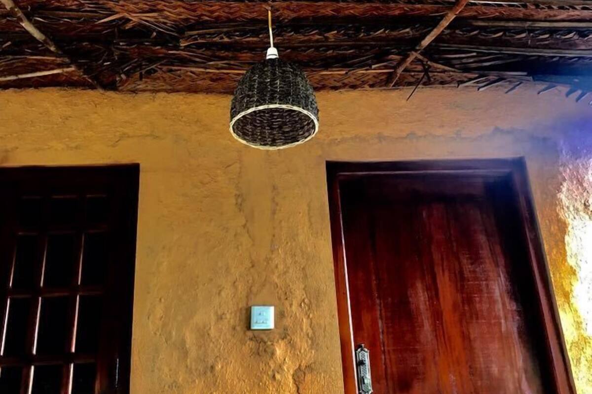 A room with a thatched roof, a woven light fixture, a wooden door, and a window with bars.