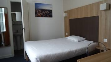 A hotel room with a single bed, a wooden headboard, a small bedside table, a mirror, and a view of a cityscape through the window.