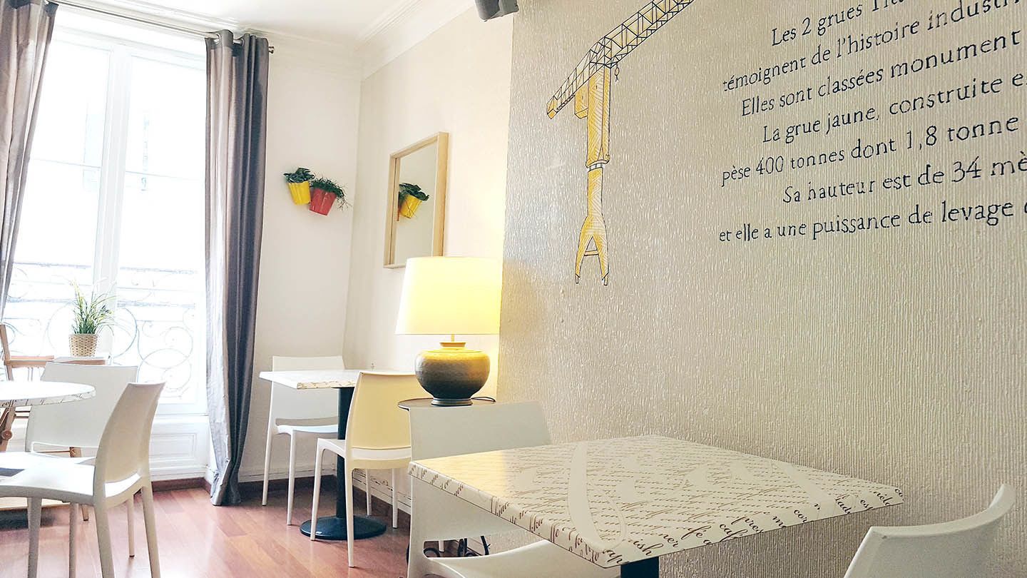 A hotel room with a dining area, a large window with curtains, a table with chairs, and a wall with a large yellow crane illustration.