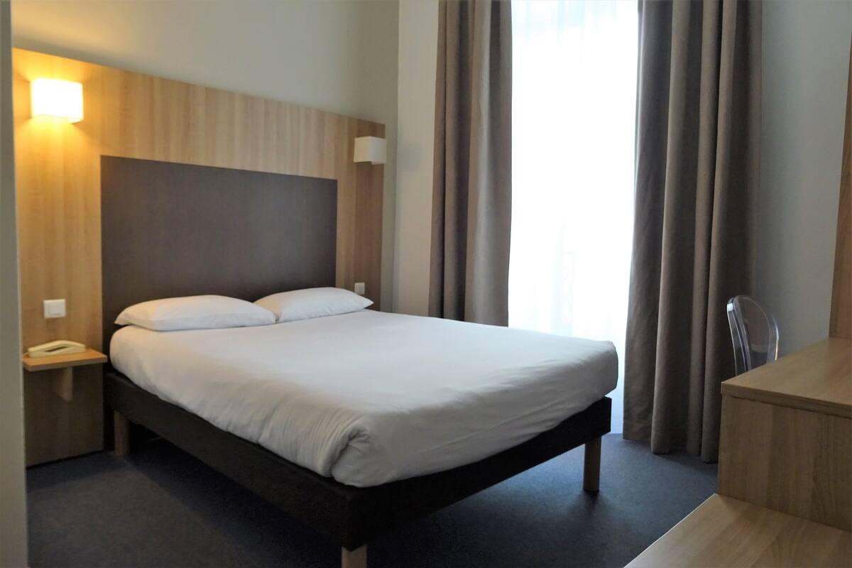 Hotellihuone, jossa on sänky, yöpöytä, lamppu ja työpöytä tuolilla.