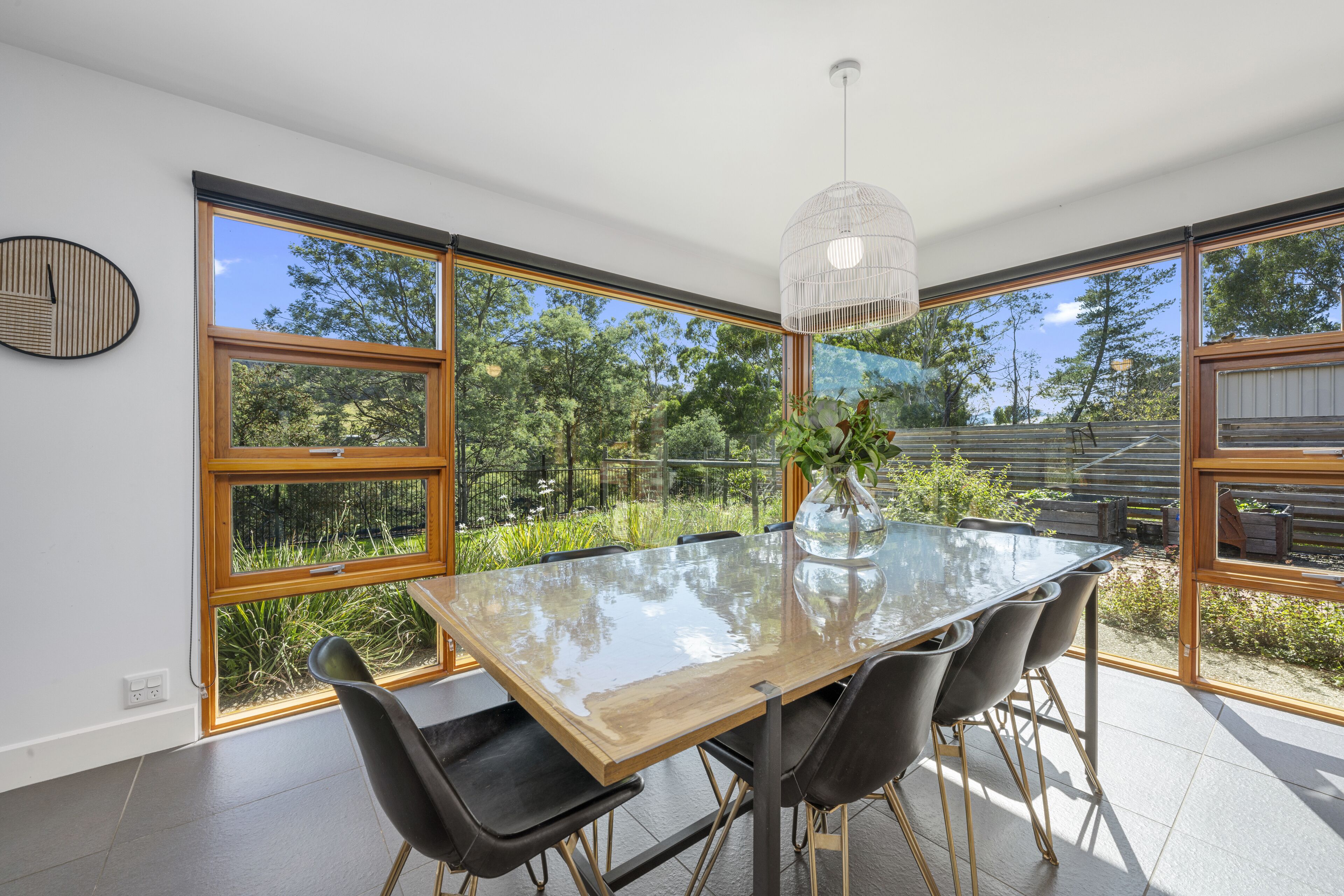 A modern dining area with a glass table, black chairs, and a view of the outdoors through large windows.