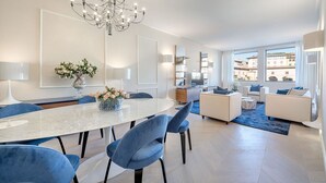 A modern living room with a dining area, a chandelier, and a view of buildings through large windows. - Luxury Blue River by Mmega (Firenze)