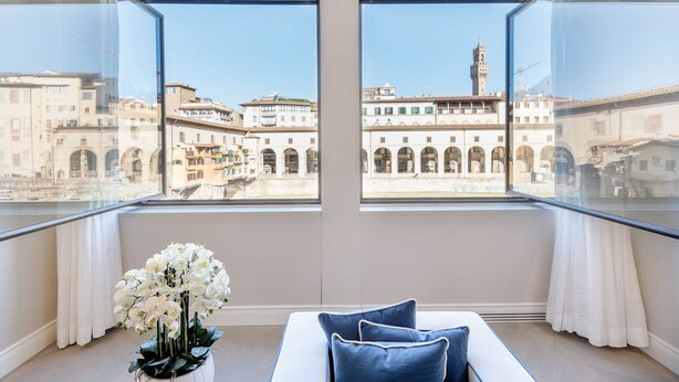 A room with a bed, a vase of flowers, and large windows with a view of historic buildings. - Luxury Blue River by Mmega (Firenze)