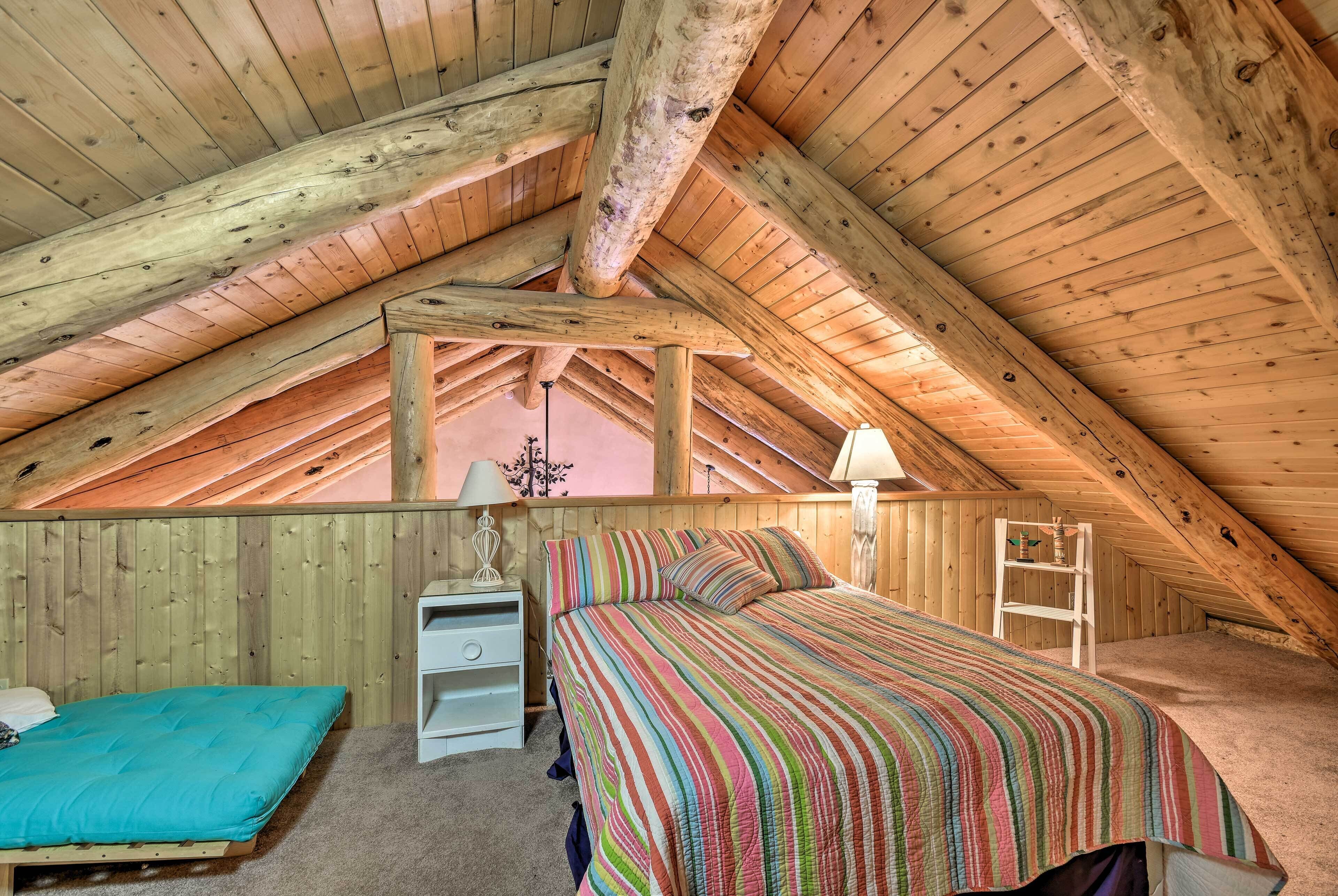 A cozy attic bedroom with a wooden ceiling, a bed with a colorful striped bedspread, a small white nightstand, and a ladder.