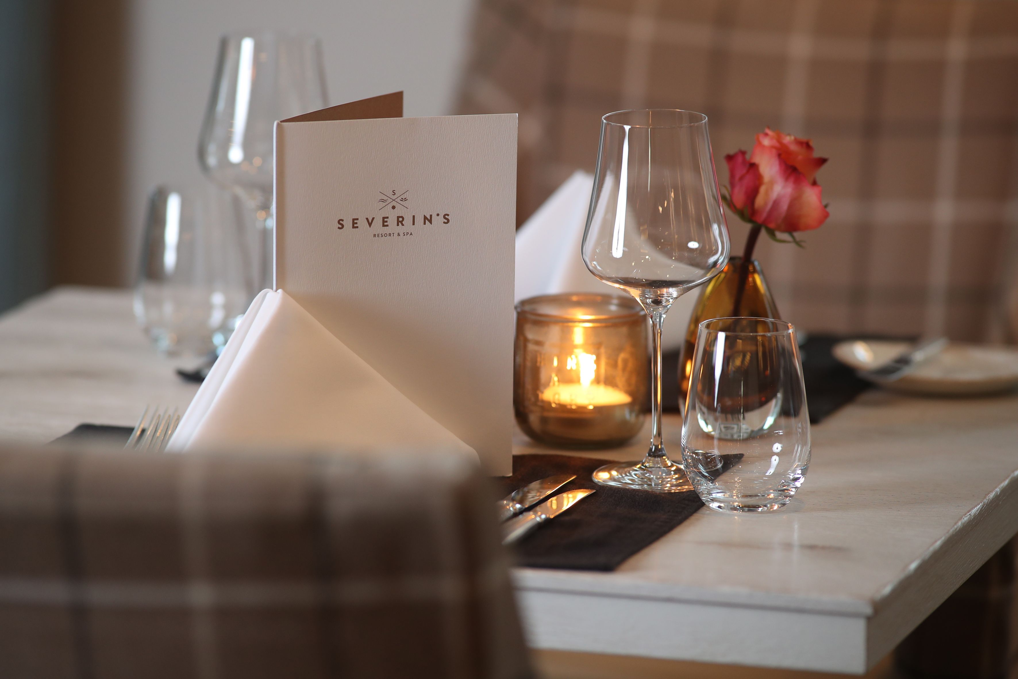 A table set with a menu card, wine glasses, a candle, and a vase with a flower.