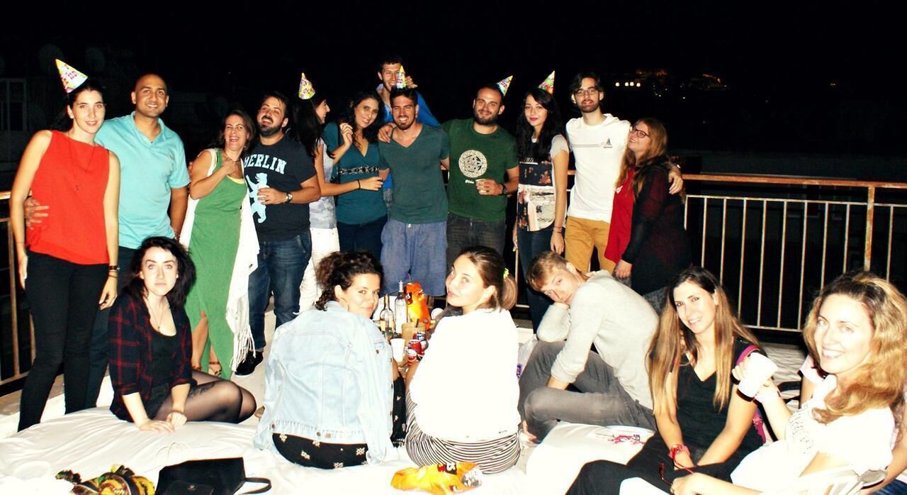 A group of people gathered around a table with drinks and snacks, seated on a bed in a room with a railing and a view of the outside.