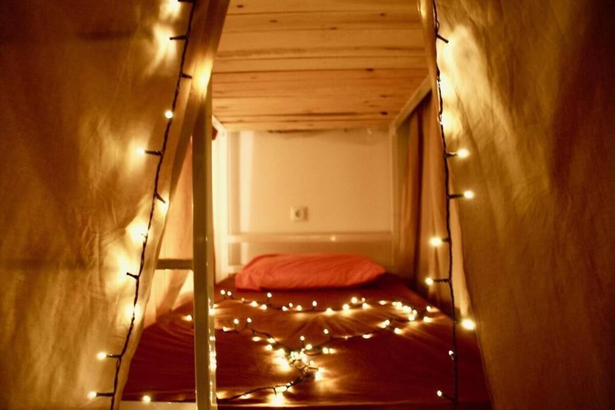 Una cama litera de madera estrecha con un cojín rojo y luces colgantes.