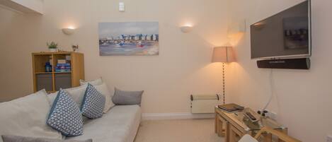 A cozy living room with a sofa, a glass coffee table, a TV mounted on the wall, and a painting above the sofa.