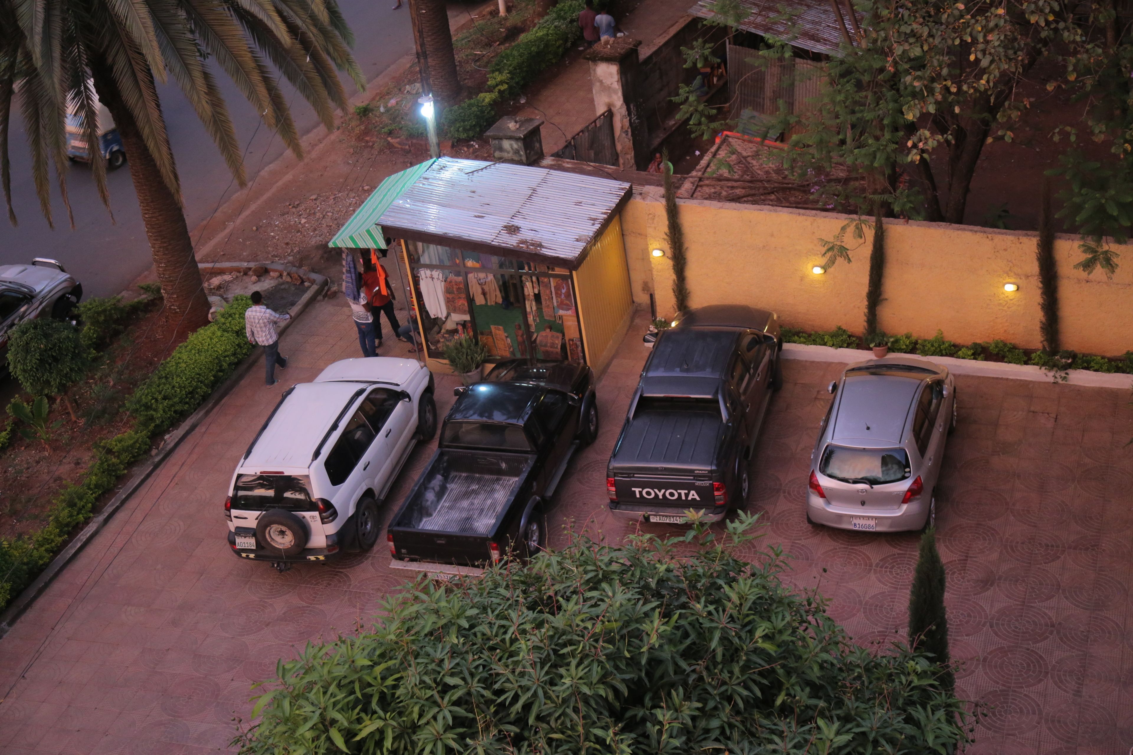 A parking area with several cars, including a Toyota pickup truck, and a small shop with a green awning.