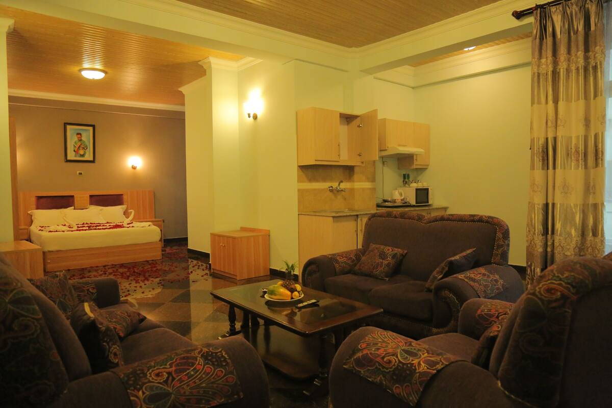 A living room with a sofa set, a coffee table, a bed with a floral bedspread, and a kitchen area with cabinets and a microwave.