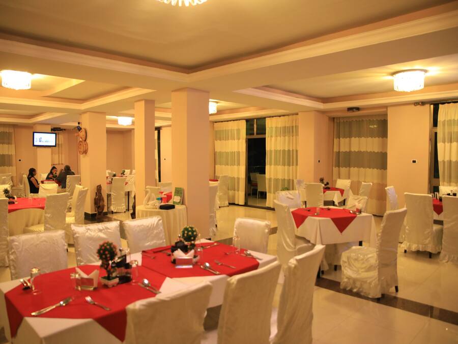 Un salón de banquetes con mesas dispuestas para un evento formal, sillas blancas y una pantalla de televisión al fondo.