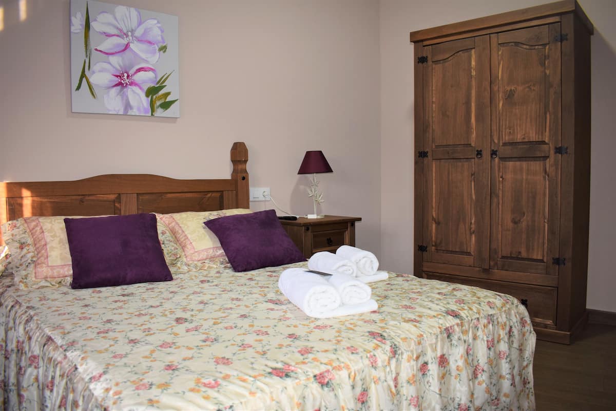 Une chambre à coucher avec une couvre-lit à motifs floraux, deux coussins violets, une armoire en bois et une table de chevet avec une lampe.