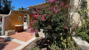 Un patio con suelo de baldosas, una mesa y sillas, rodeado de plantas florecientes y un muro de piedra.