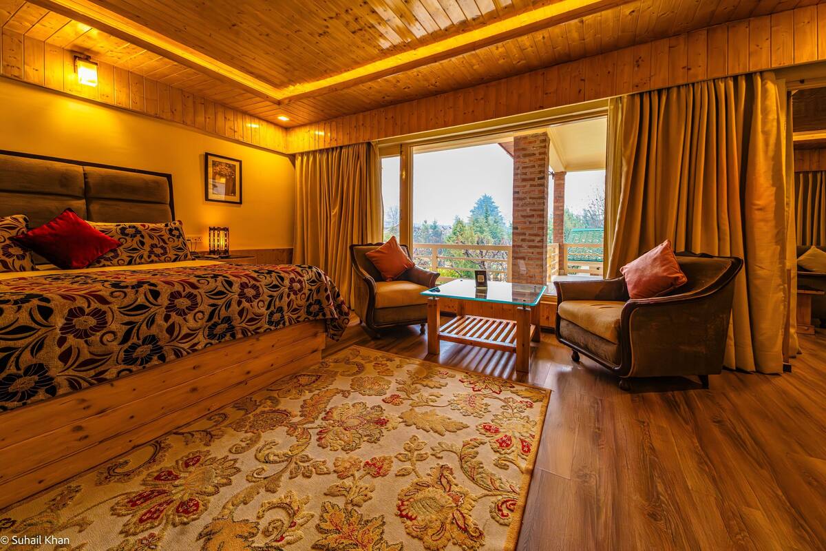 Quarto com painéis de madeira, uma cama, duas poltronas, uma mesa de centro de vidro e vista para o exterior.