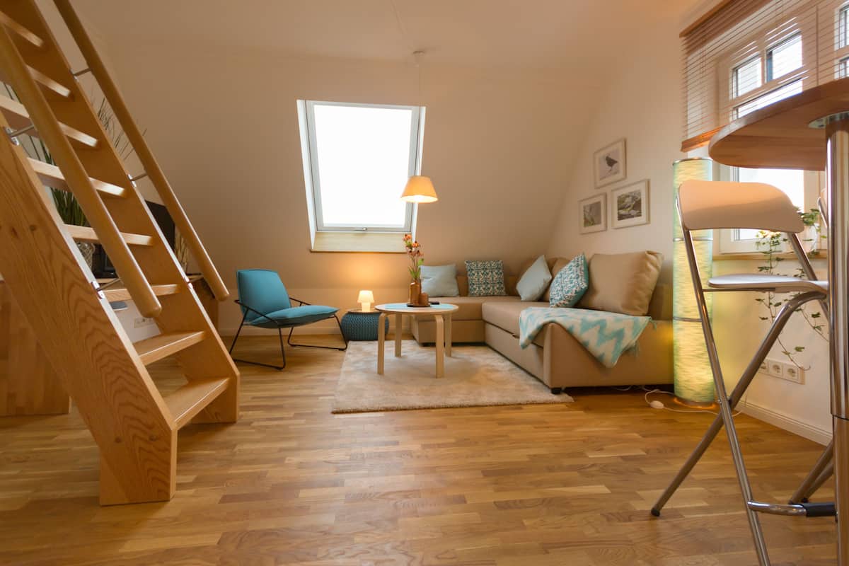 A modern living room with a wooden ladder, a blue chair, a sofa with patterned cushions, a small round table, and a floor lamp.