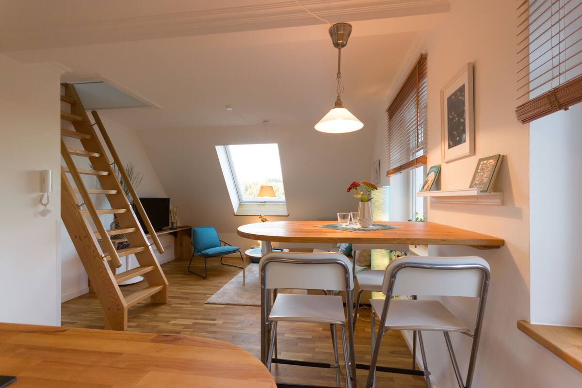 A modern kitchen with a wooden table, two metal chairs, a wooden ladder, and a window with blinds.