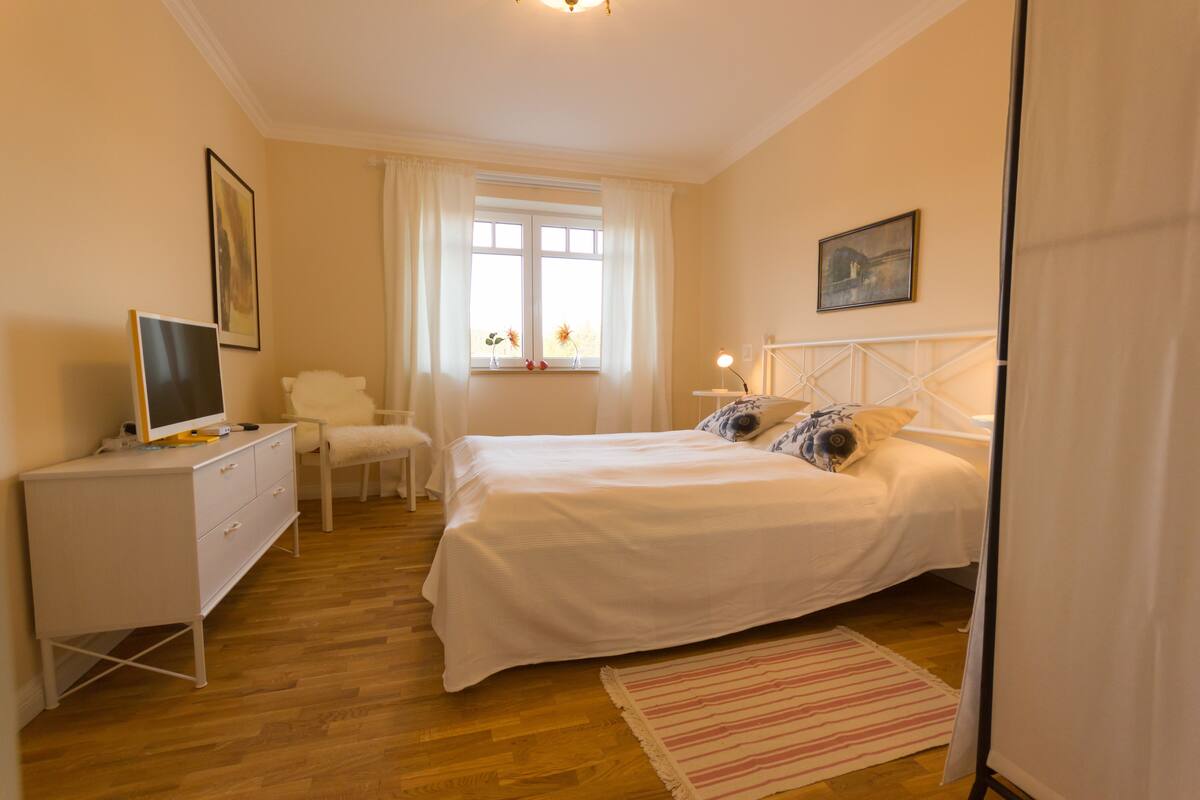 A bedroom with a bed, a desk, a chair, a window with curtains, and a bedside table.