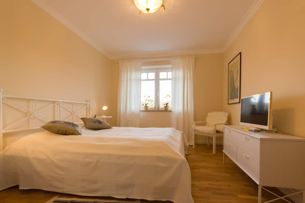 A bedroom with a bed, a TV, a chair, and a window with curtains.