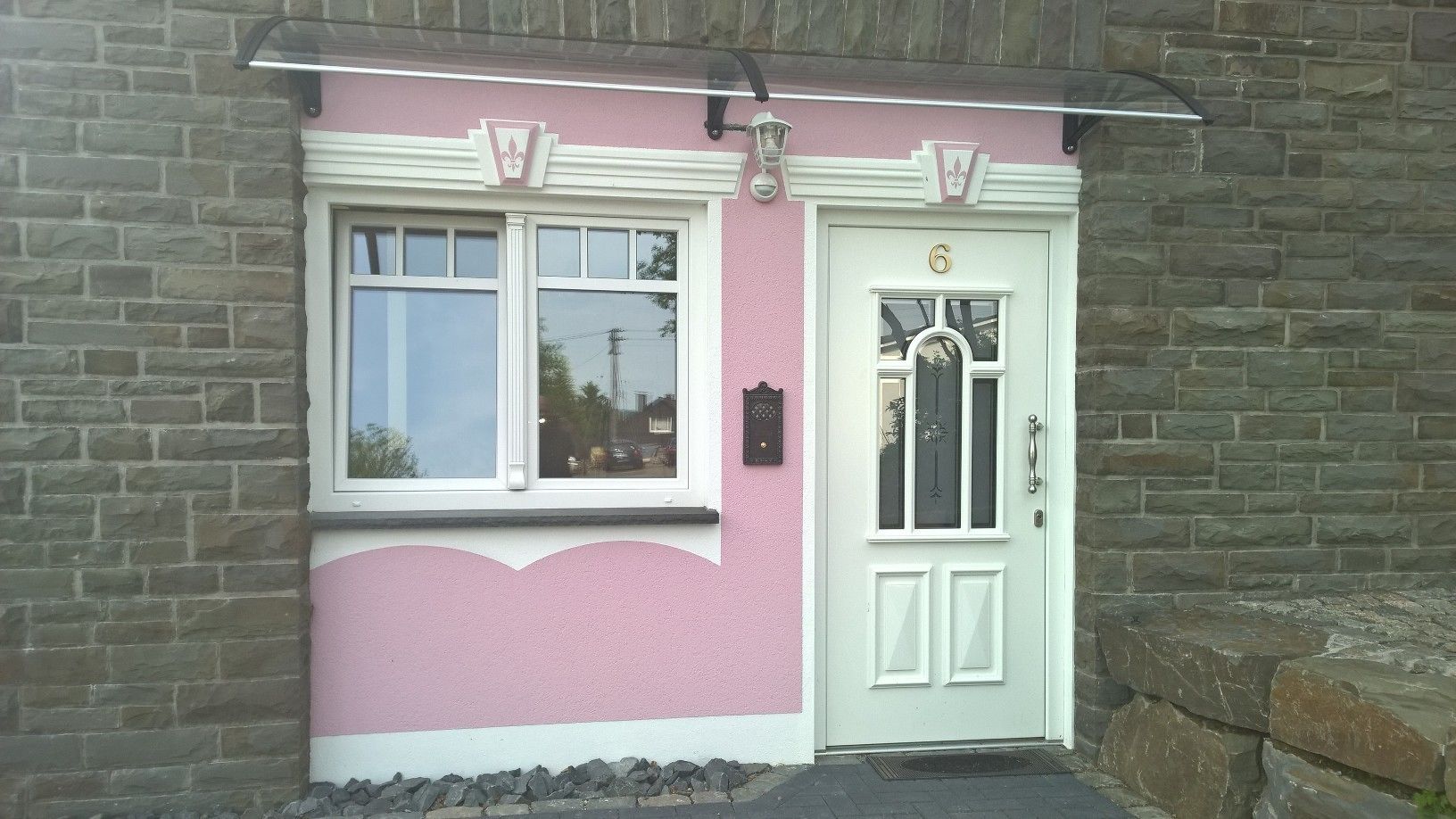 A pink building with a white door and window, a stone wall, and a small step leading to the entrance.