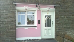 A pink building with a white door and window, a stone wall, and a small step leading to the entrance. - Villa Ruth (Reichshof)