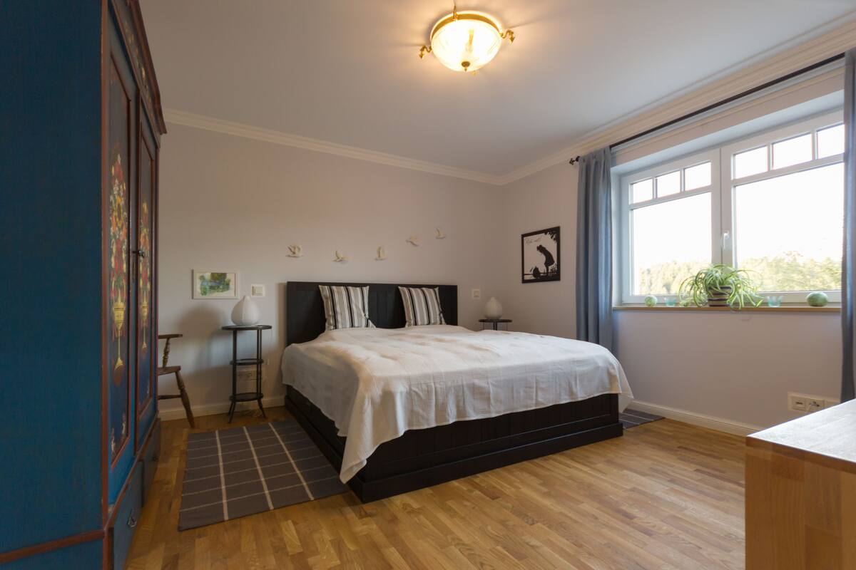 A bedroom with a bed, a nightstand, a lamp, a window with curtains, and a view of greenery outside.