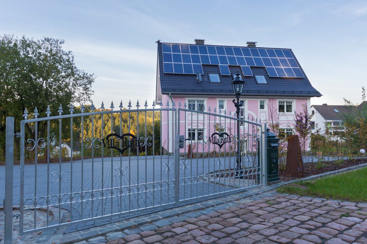Une entrée fermée par un portail mène à une maison rose, dont le toit est équipé de panneaux solaires, et le portail lui-même est en métal et agrémenté d’éléments décoratifs.