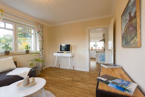 A modern living room with a flat-screen TV on a white stand, a sofa, a coffee table with a candle and a decorative item, a wooden dining table with magazines, and a view of the kitchen through an open door. - Villa Ruth (Reichshof)