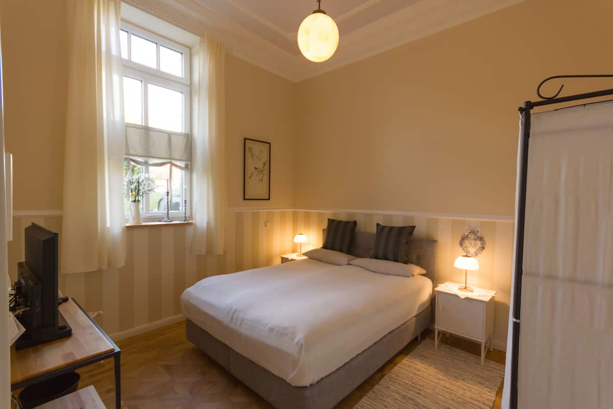 A hotel room with a large bed, bedside tables with lamps, a window with curtains, and a television on a desk.