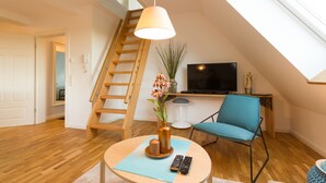 A modern living room with a wooden staircase, a round table with a vase and remote controls, a blue chair, and a flat-screen TV on a stand. - Villa Ruth (Reichshof)